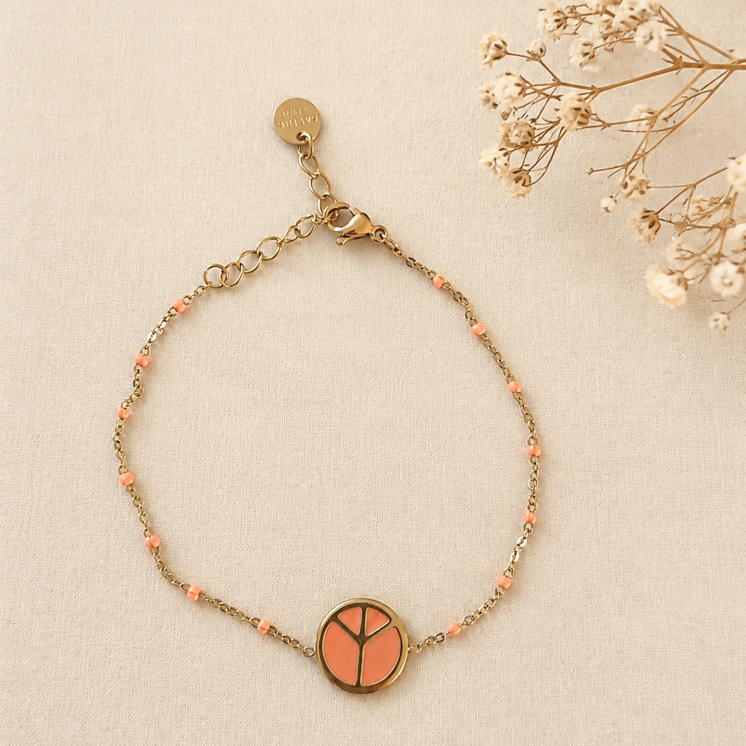 Bracelet fin doré avec médaille géométrique émaillée corail et petites perles corail, bijou élégant porté au poignet sur fond en lin clair.