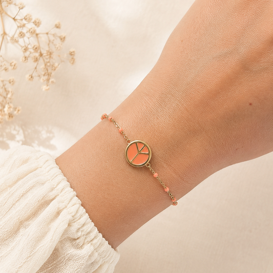 Bracelet doré en acier inoxydable avec médaille émaillée corail et petites perles corail porté au poignet.